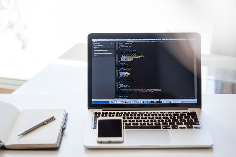 A close-up of a clean workspace featuring an open laptop displaying lines of computer code on a dark background, positioned on a white desk. To the left of the laptop, there is an open white notebook 