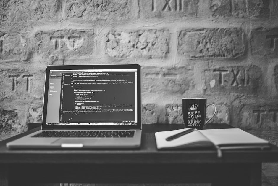 A close-up of a clean workspace featuring an open laptop displaying lines of computer code on a dark background, positioned on a white desk. To the left of the laptop, there is an open white notebook 